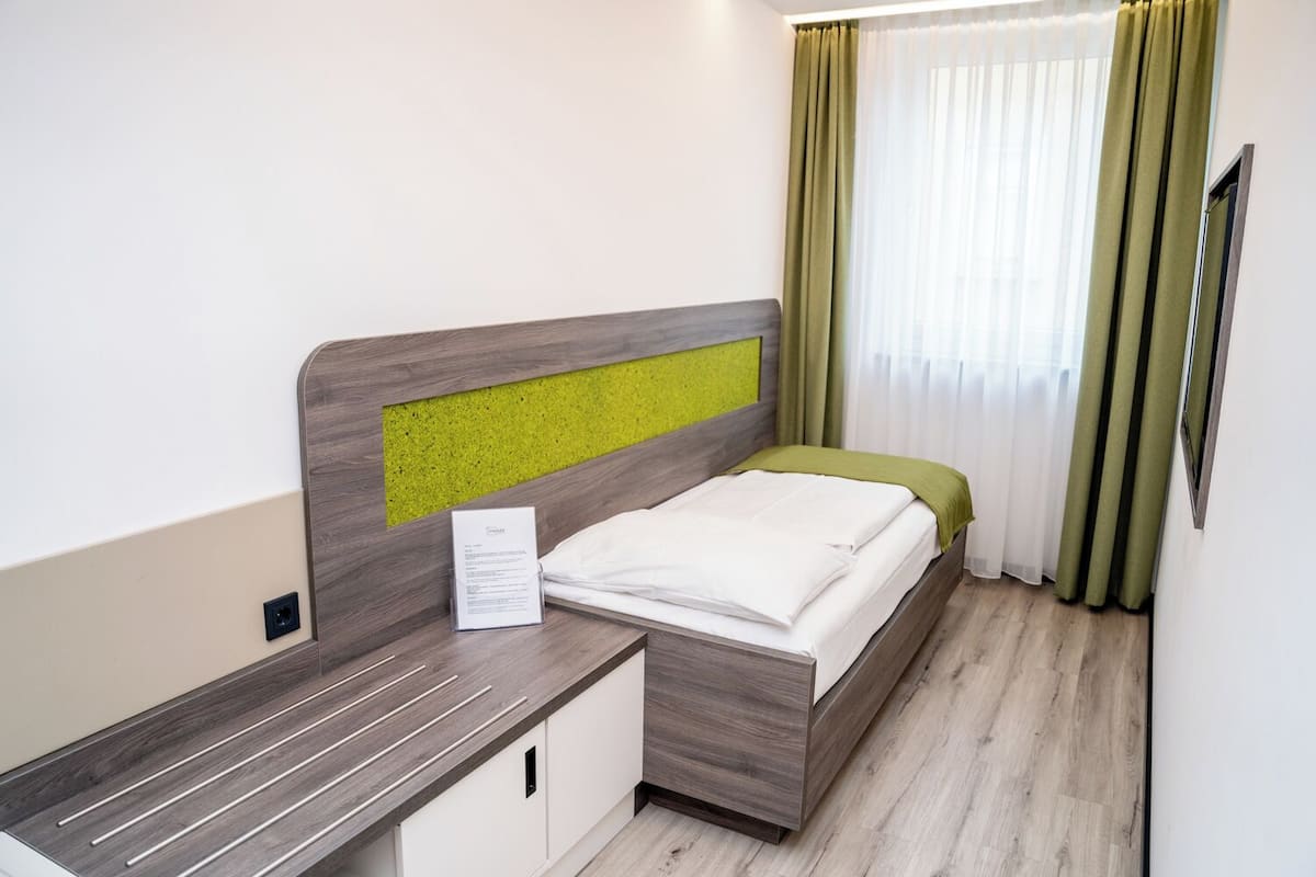 Ein ordentlich bezogenes Bett mit einem grünen Kopfteil, ein Nachttisch mit einer Lampe und ein Fenster mit grünen Vorhängen.
