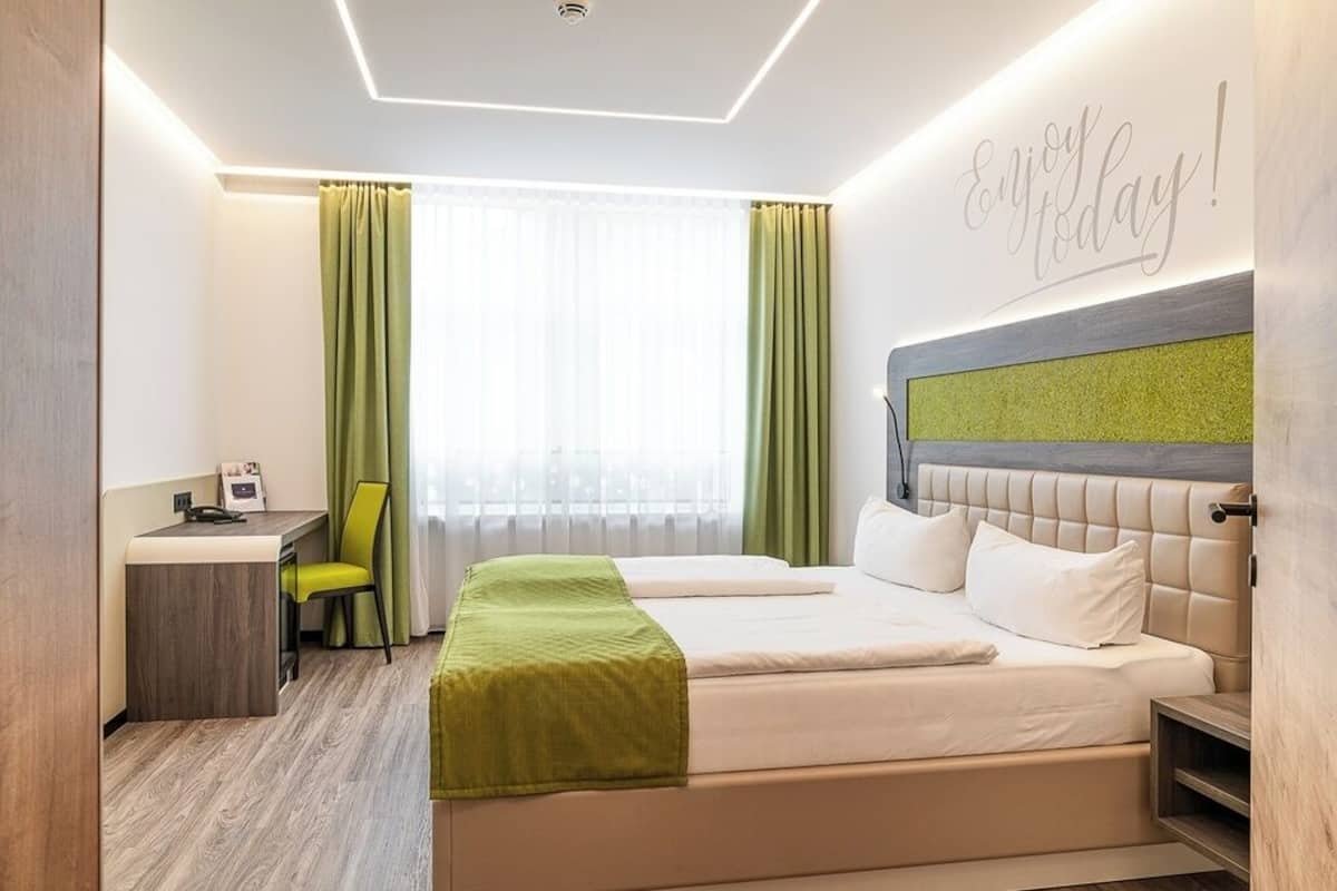 Ein Hotelzimmer mit Bett, Schreibtisch und Stuhl. Ein Fenster mit Vorhängen, eine Wand mit einem motivierenden Zitat und einer Deckenleuchte.