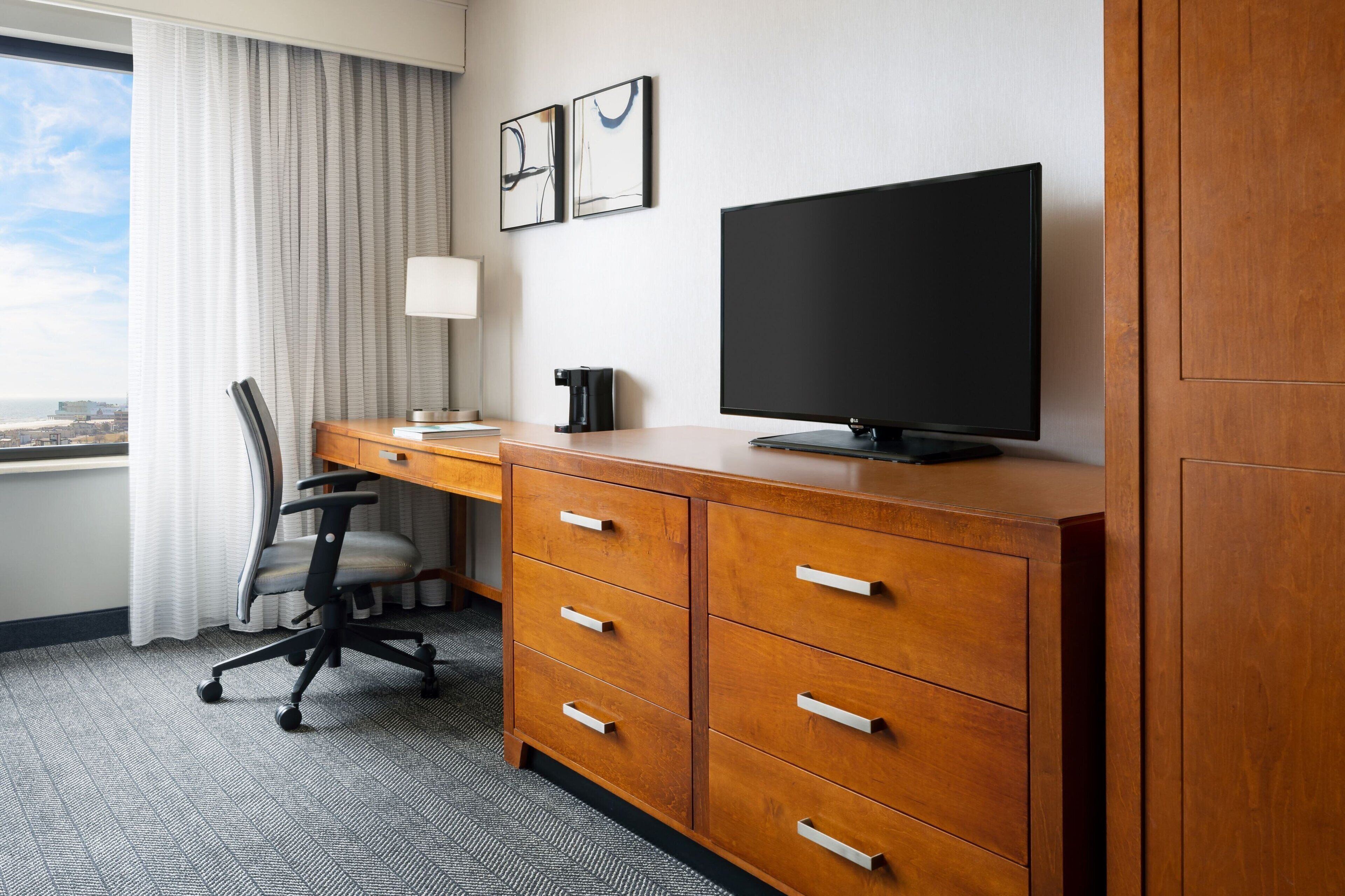 A hotel room with a wooden desk, a flat-screen TV, a chair, a lamp, and a window with curtains.