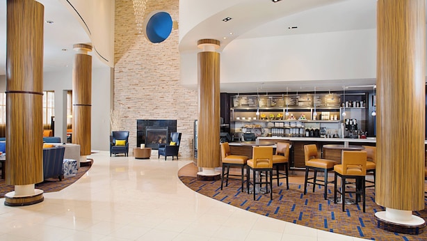 Un vestíbulo moderno con altas columnas de madera, una pared de piedra, una zona de bar y diferentes disposiciones de asientos.