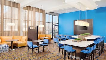 Un hall d’hôtel avec un mur d’accent bleu, un espace salon avec un canapé et des fauteuils, un coin bar avec des tabourets hauts et une grande fenêtre laissant entrer la lumière naturelle.