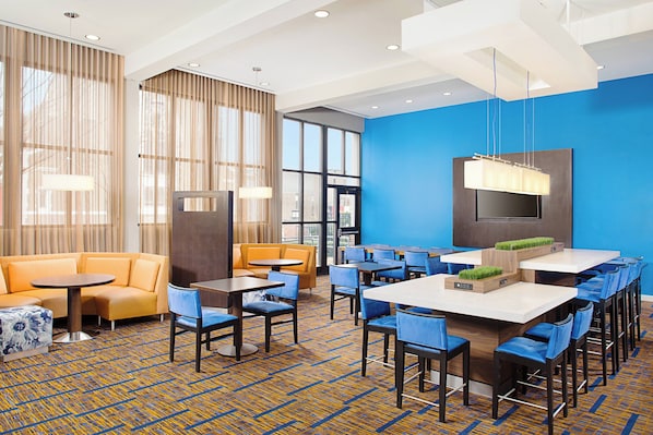 A hotel lobby with a blue accent wall, a seating area with a sofa and chairs, a bar area with high stools, and a large window providing natural light.