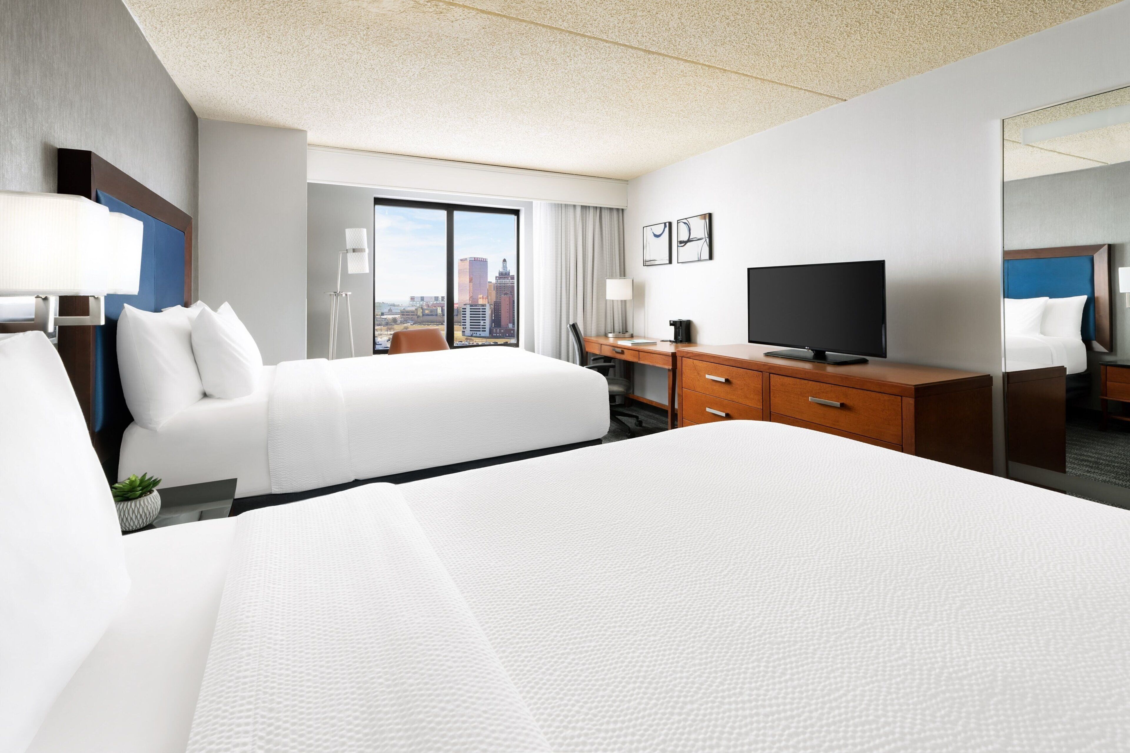 Habitación de hotel con una cama grande, un escritorio con televisor y vista a la ciudad desde la ventana.