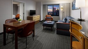 Habitación de hotel con zona de comedor, sofá, televisor y ventana con vista a la ciudad.