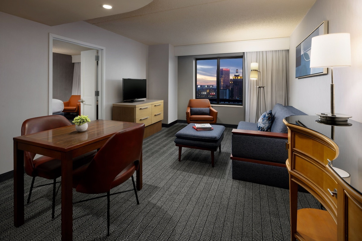 Habitación de hotel con zona de comedor, sofá, televisor y ventana con vista a la ciudad.