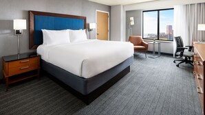 Habitación de hotel con una cama grande, un escritorio, una silla y vistas a la ciudad a través de un ventanal amplio.