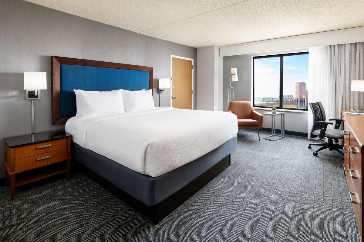 Ein Hotelzimmer mit einem großen Bett, einem Schreibtisch, einem Sessel und Blick auf die Stadt durch ein großes Fenster.