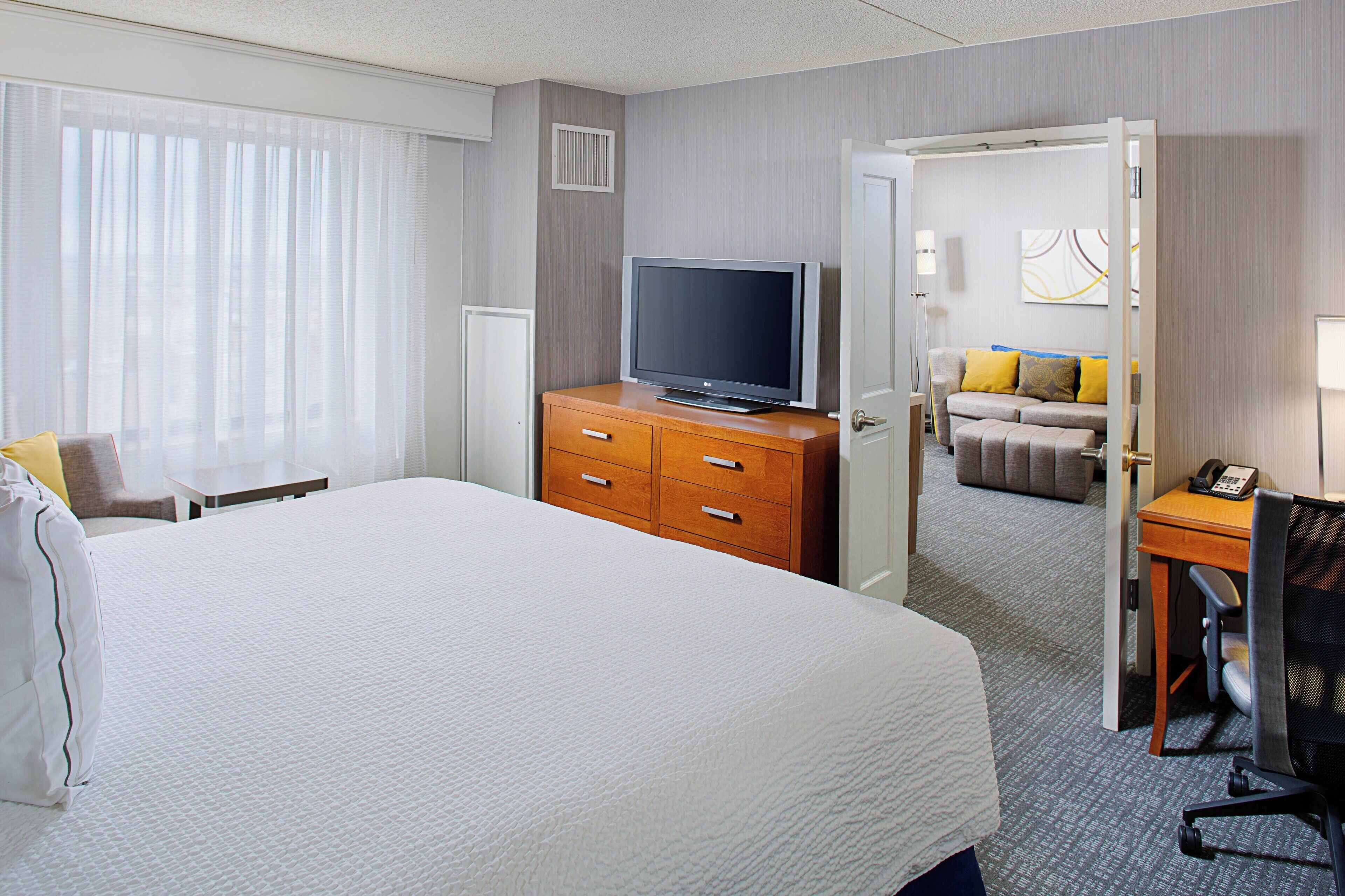 A hotel room with a large bed, a wooden dresser with a flat-screen TV, a desk with a chair, and a sofa in the background.