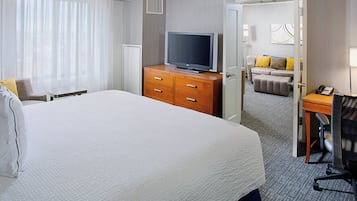 A hotel room with a large bed, a wooden dresser with a flat-screen TV, a desk with a chair, and a sofa in the background.