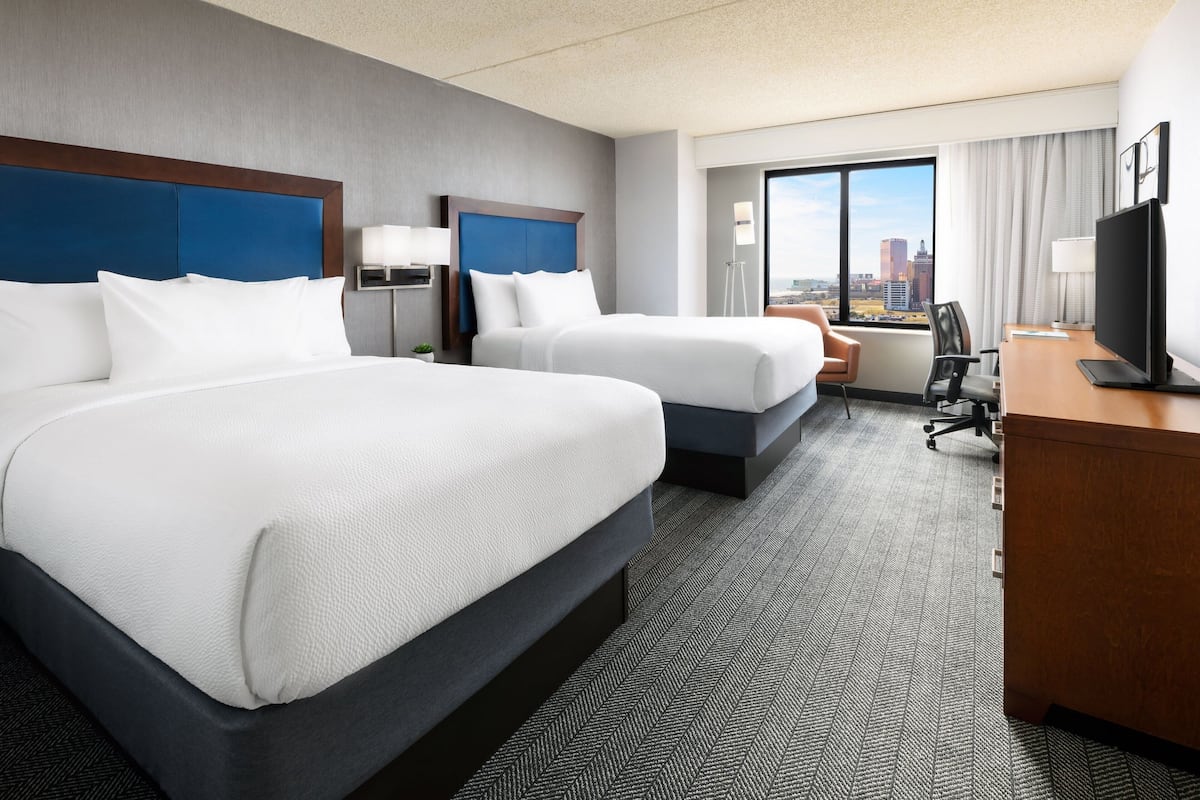 Ein Hotelzimmer mit zwei Betten, einem Schreibtisch, einem Sessel, einem Fernseher und einem großen Fenster mit Blick auf die Stadt.