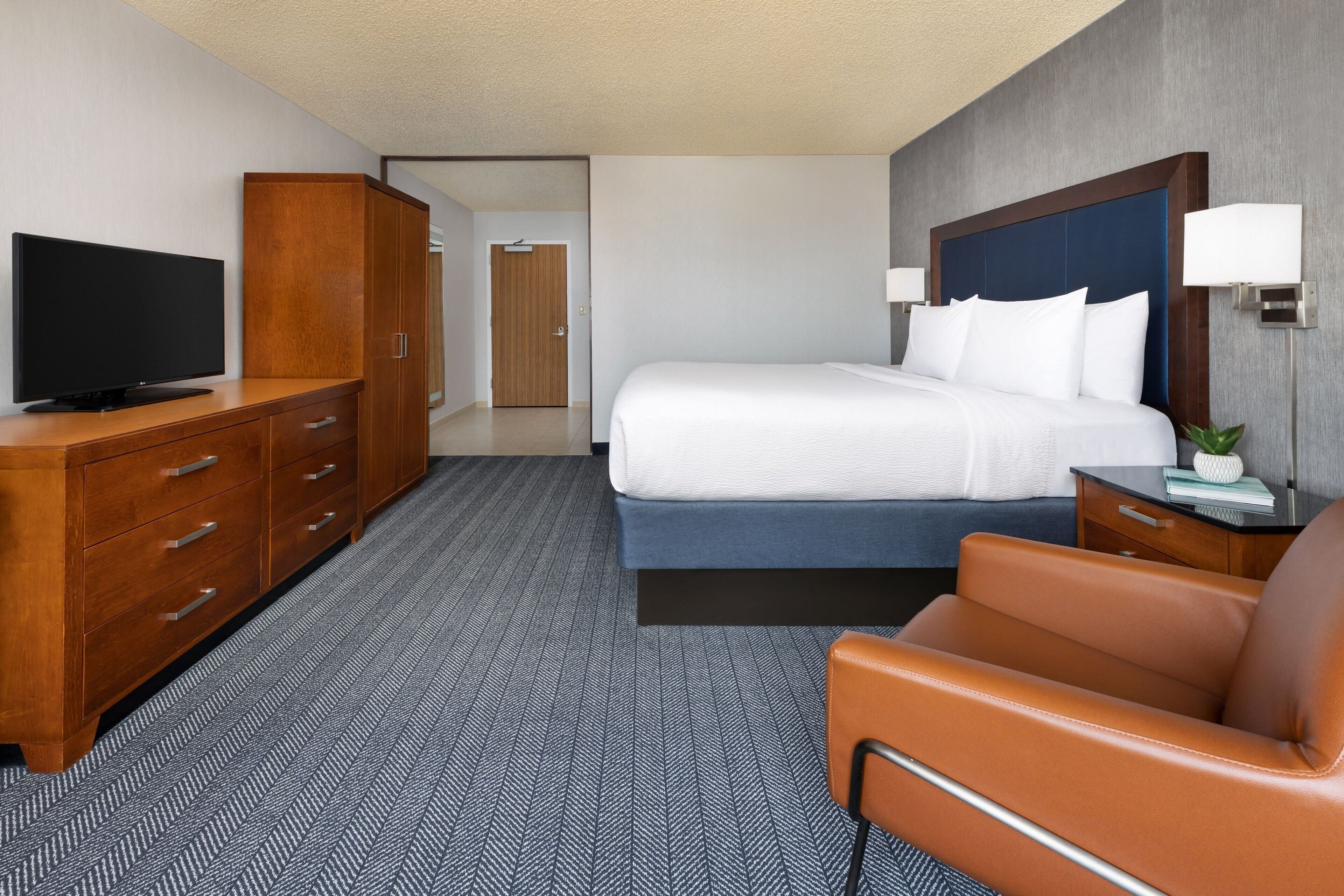 Habitación de hotel con una cama, un televisor sobre una cómoda de madera, un escritorio con lámpara y una silla.