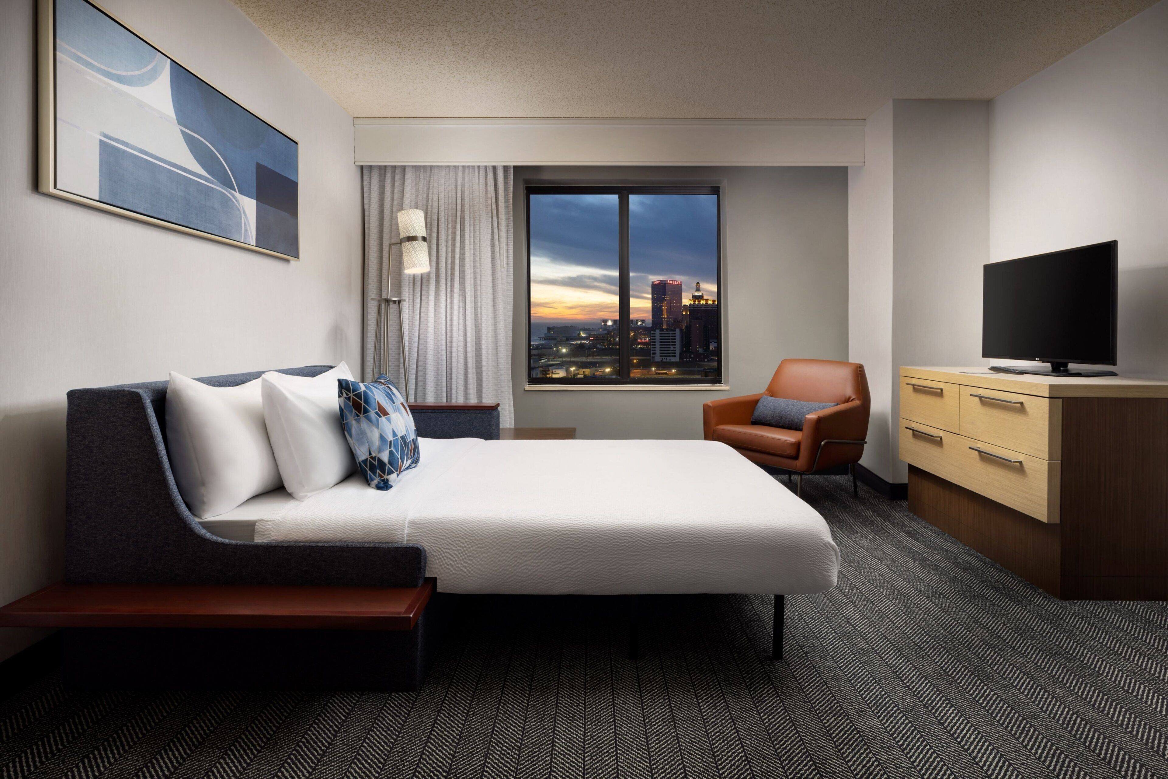 Habitación de hotel con cama, escritorio, silla, televisión y ventana con vista a la ciudad.