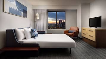 A hotel room with a bed, a desk, a chair, a television, and a window with a city view.
