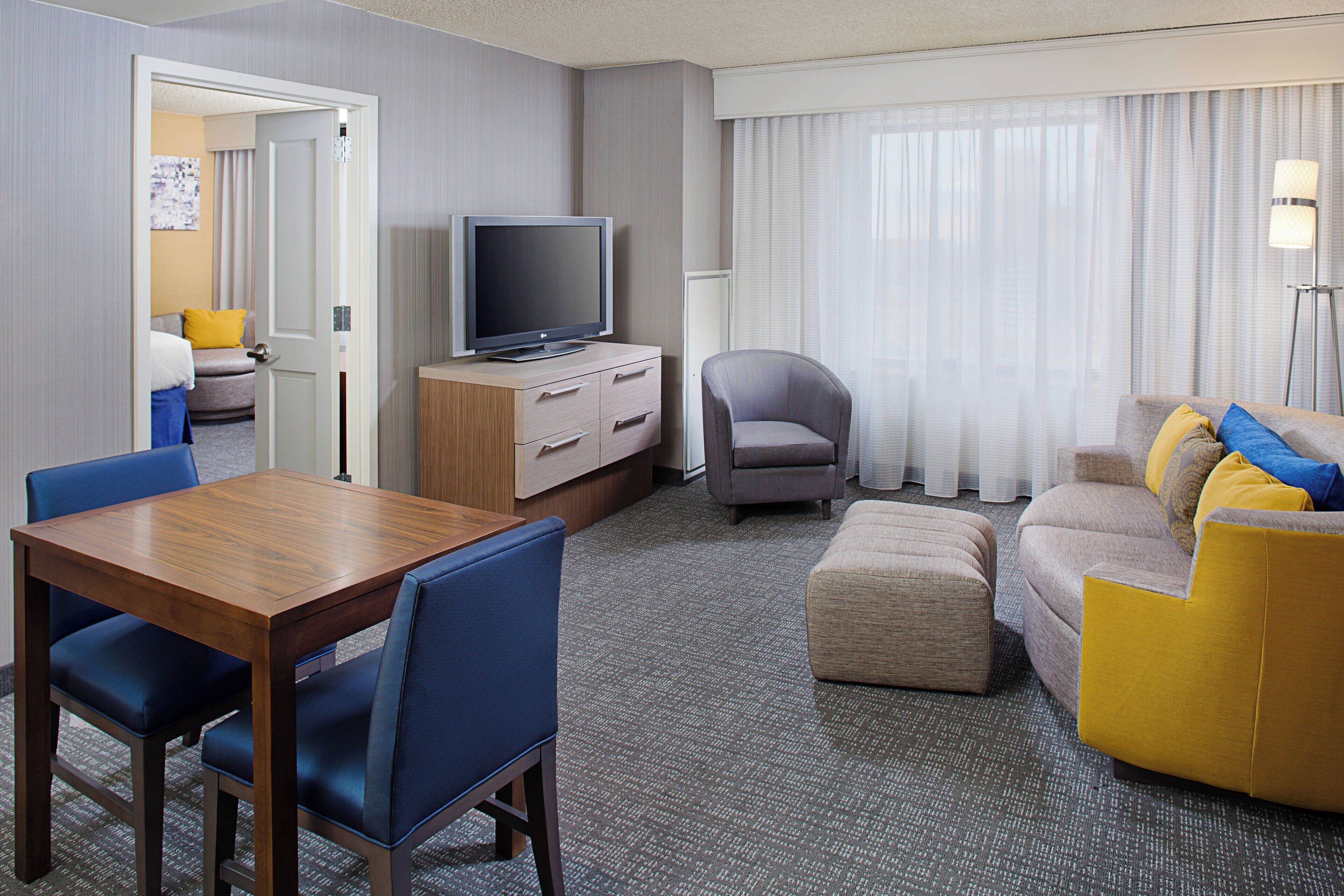 Habitación de hotel con una mesa de comedor de madera, sillas azules, un sofá gris con cojines amarillos y azules, un sillón gris, un televisor de pantalla plana sobre un mueble de madera y un gran ventanal con cortinas translúcidas.