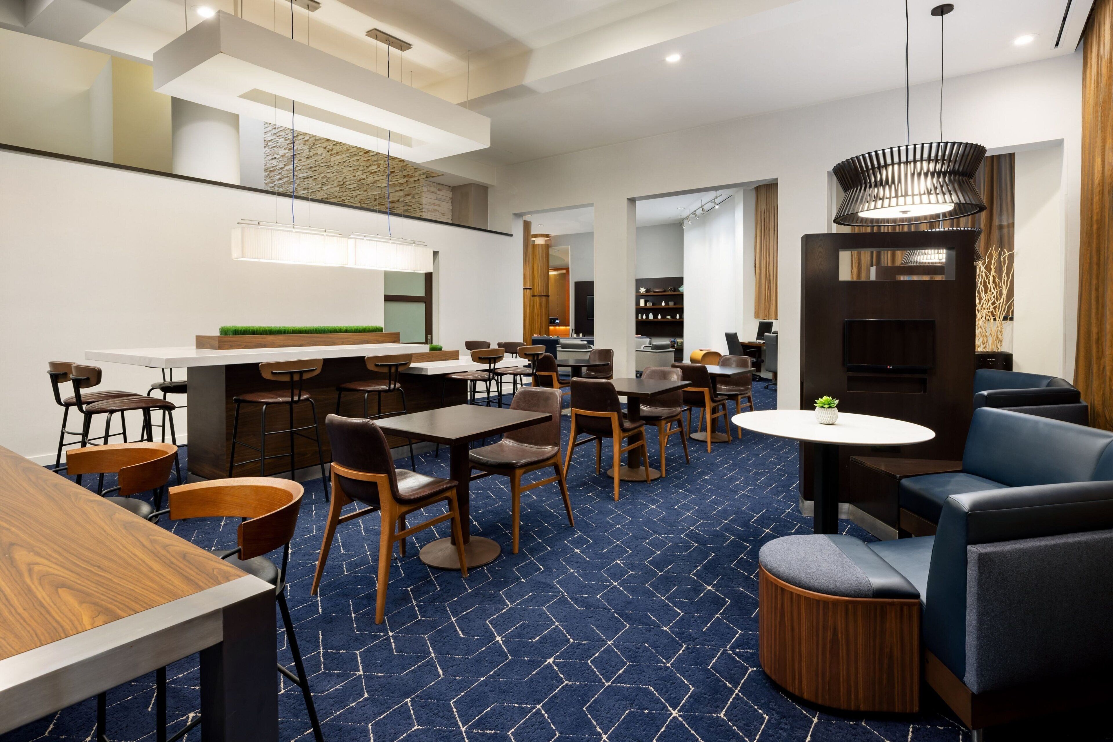 A modern hotel lobby with a blue patterned carpet, wooden tables and chairs, a bar area, and a seating area with sofas and a TV.