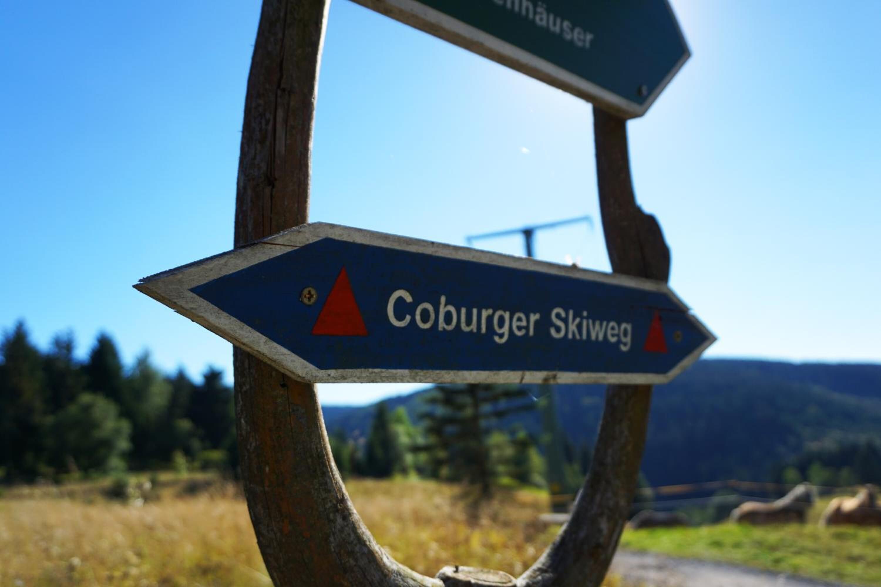 A directional signpost with 'Coburger Skiweg' written on it, set against a backdrop of a clear sky and a mountainous landscape.