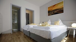 Habitación de hotel con cama doble, mesita de noche, zona de ducha y un cuadro de paisaje urbano en la pared.