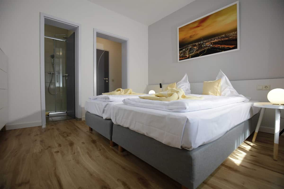 Habitación de hotel con cama doble, mesita de noche, zona de ducha y un cuadro de paisaje urbano en la pared.