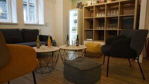 Una sala de estar con un sofá, sillas, una mesita redonda con botellas y vasos, una estantería y un refrigerador.