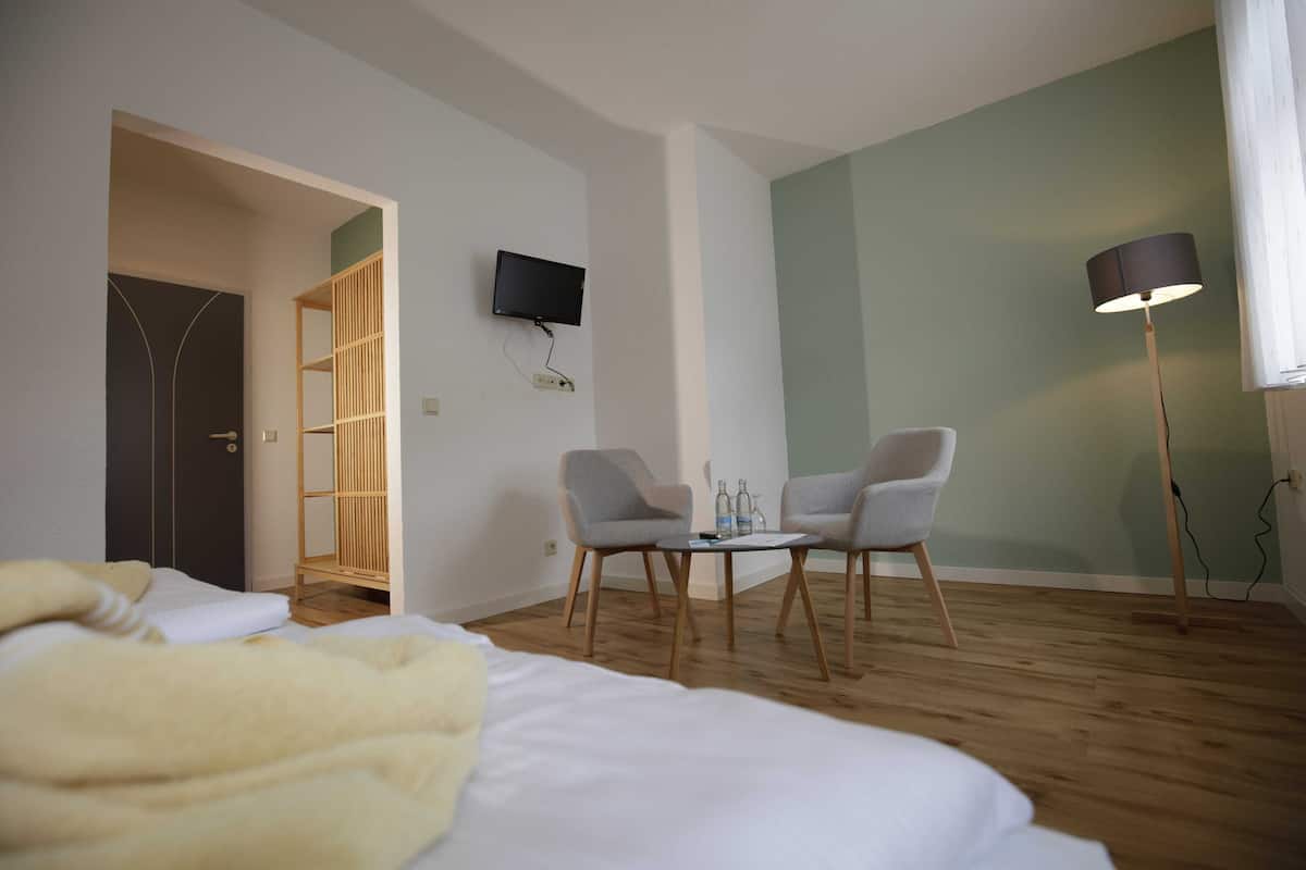Habitación de hotel con cama, dos sillas, una mesita, televisor montado en la pared y una lámpara de pie.