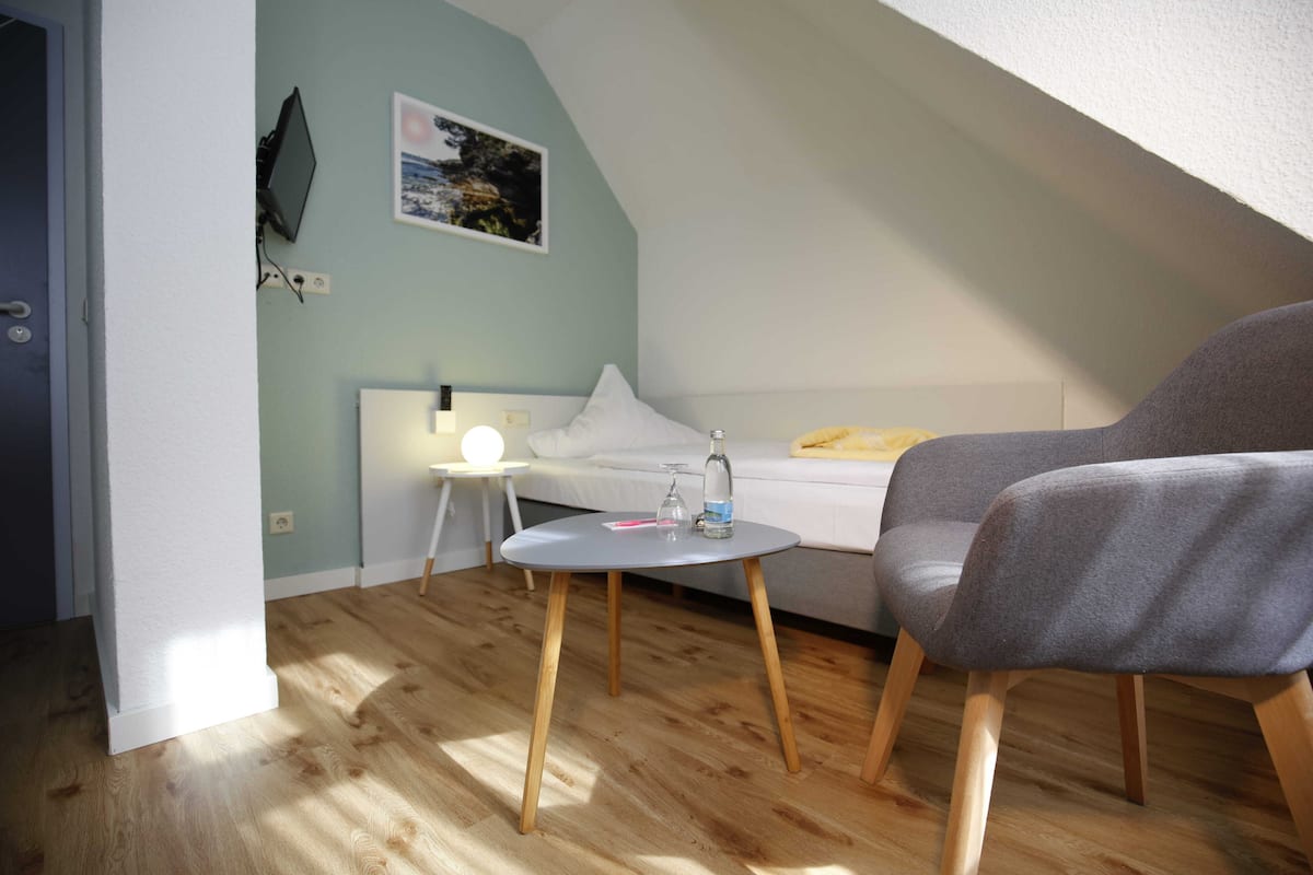 Una habitación acogedora con una cama, un sillón gris, una mesita redonda y un televisor instalado en la pared.