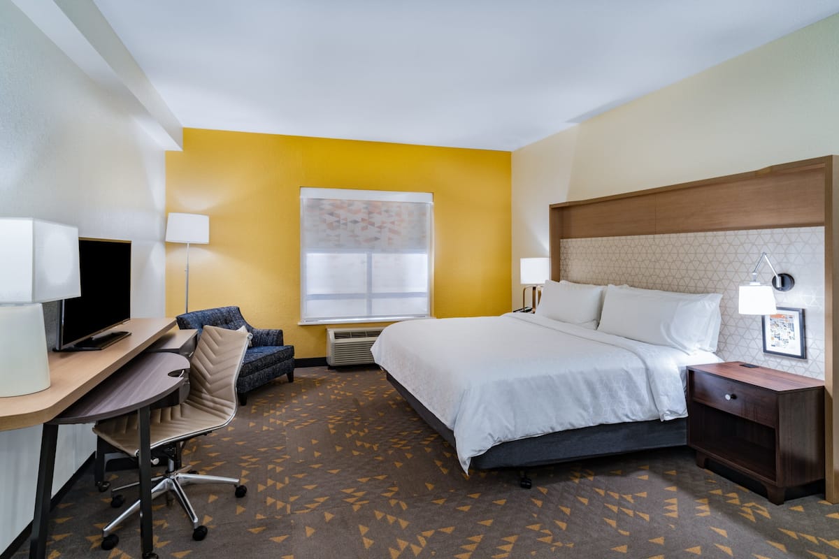 A hotel room with a bed, desk, chair, and a window with blinds.
