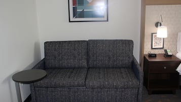 Un sofá gris con una pequeña mesa redonda a un lado, una obra de arte abstracta enmarcada en la pared, una mesita de noche con una lámpara y un teléfono.