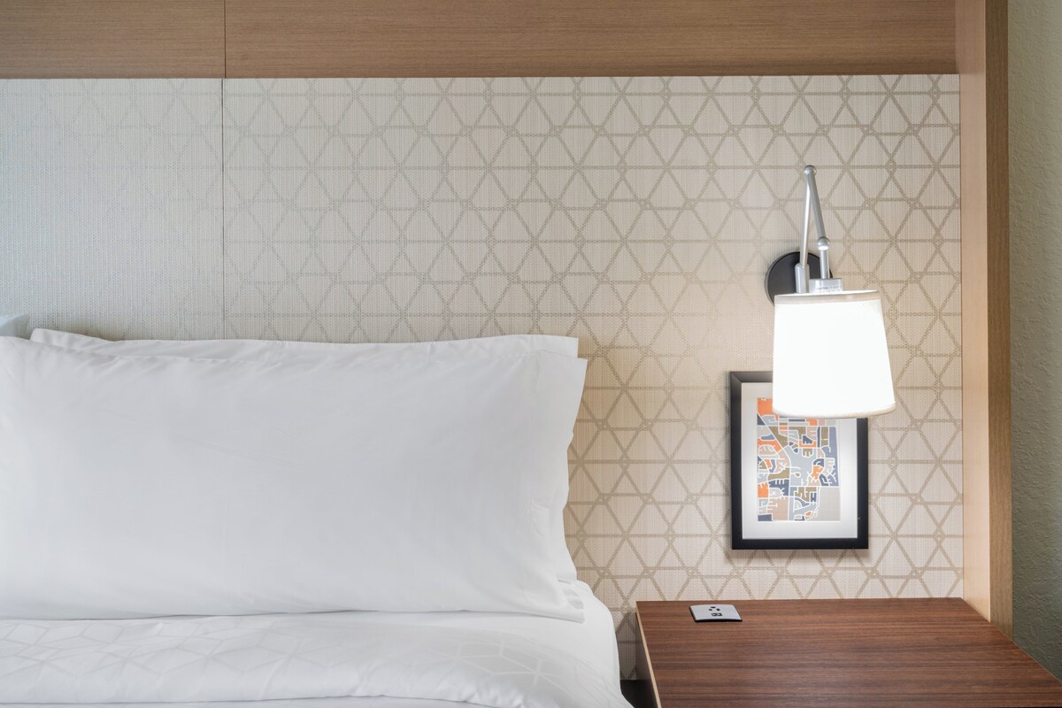 A neatly made bed with white linens, a wooden nightstand with a framed picture and a lamp, and a patterned wall.