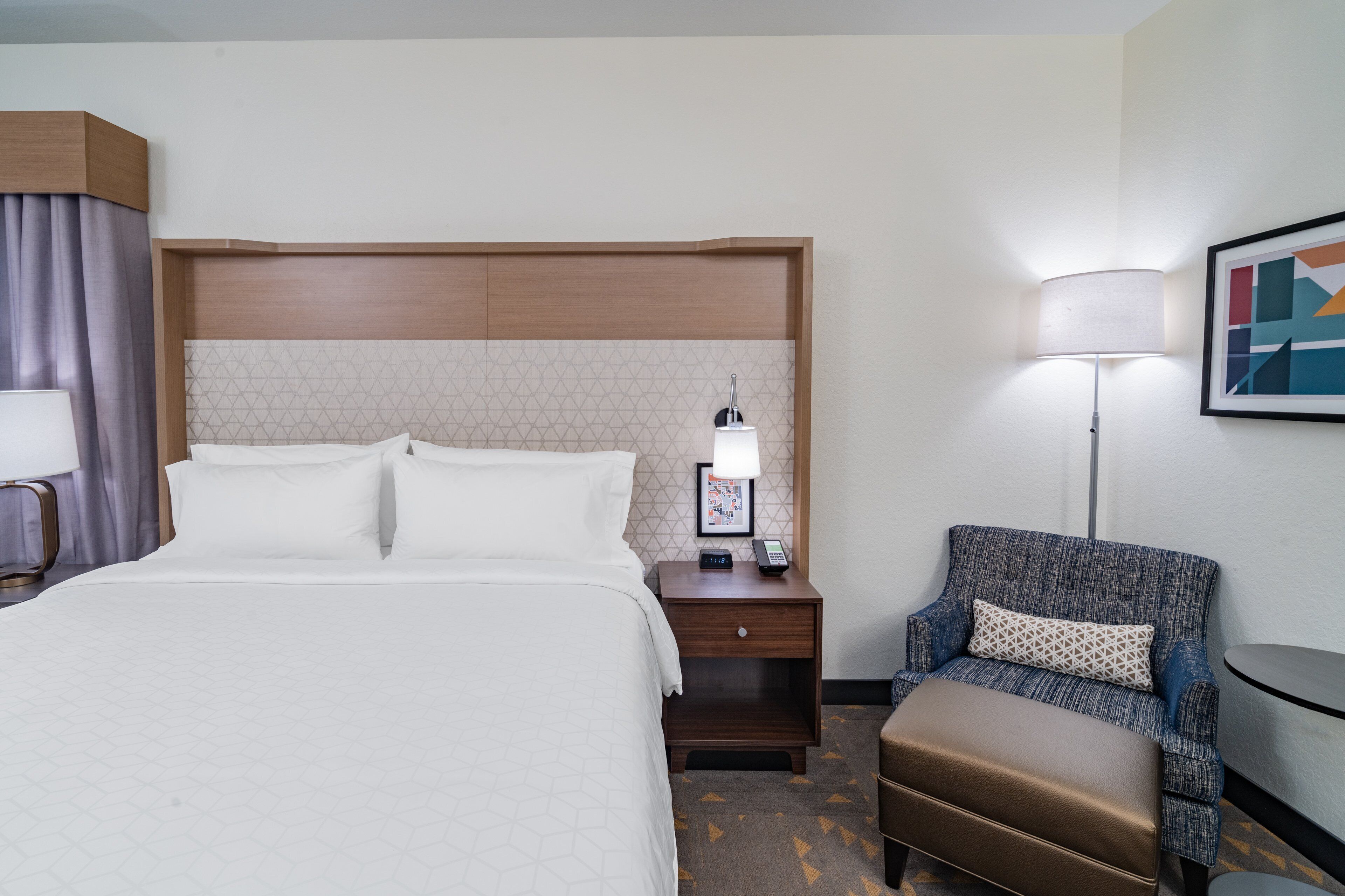 Habitación de hotel con cama, mesita de noche, lámpara, silla y un cuadro en la pared.