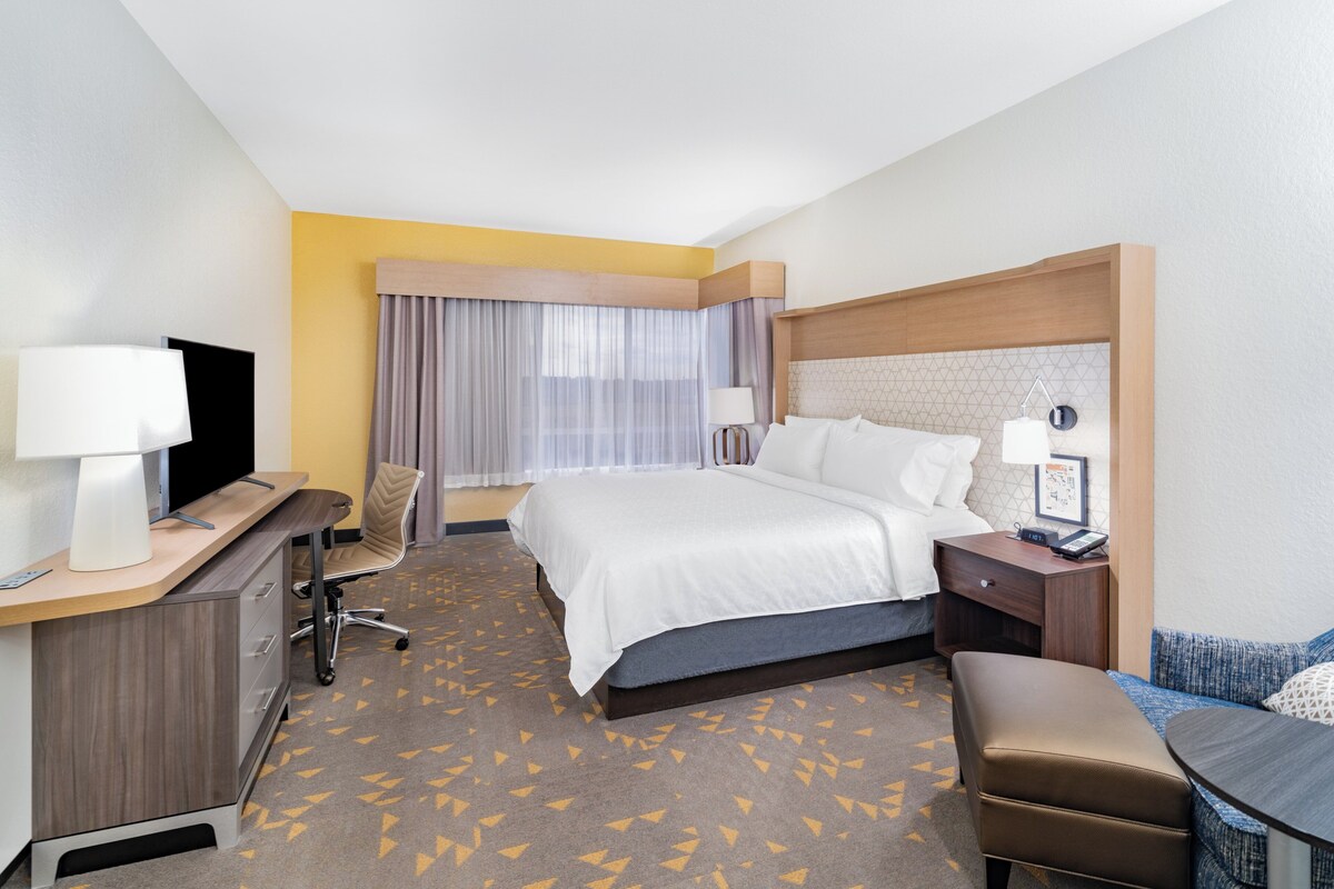 A hotel room with a bed, desk, chair, and a small table with a lamp.