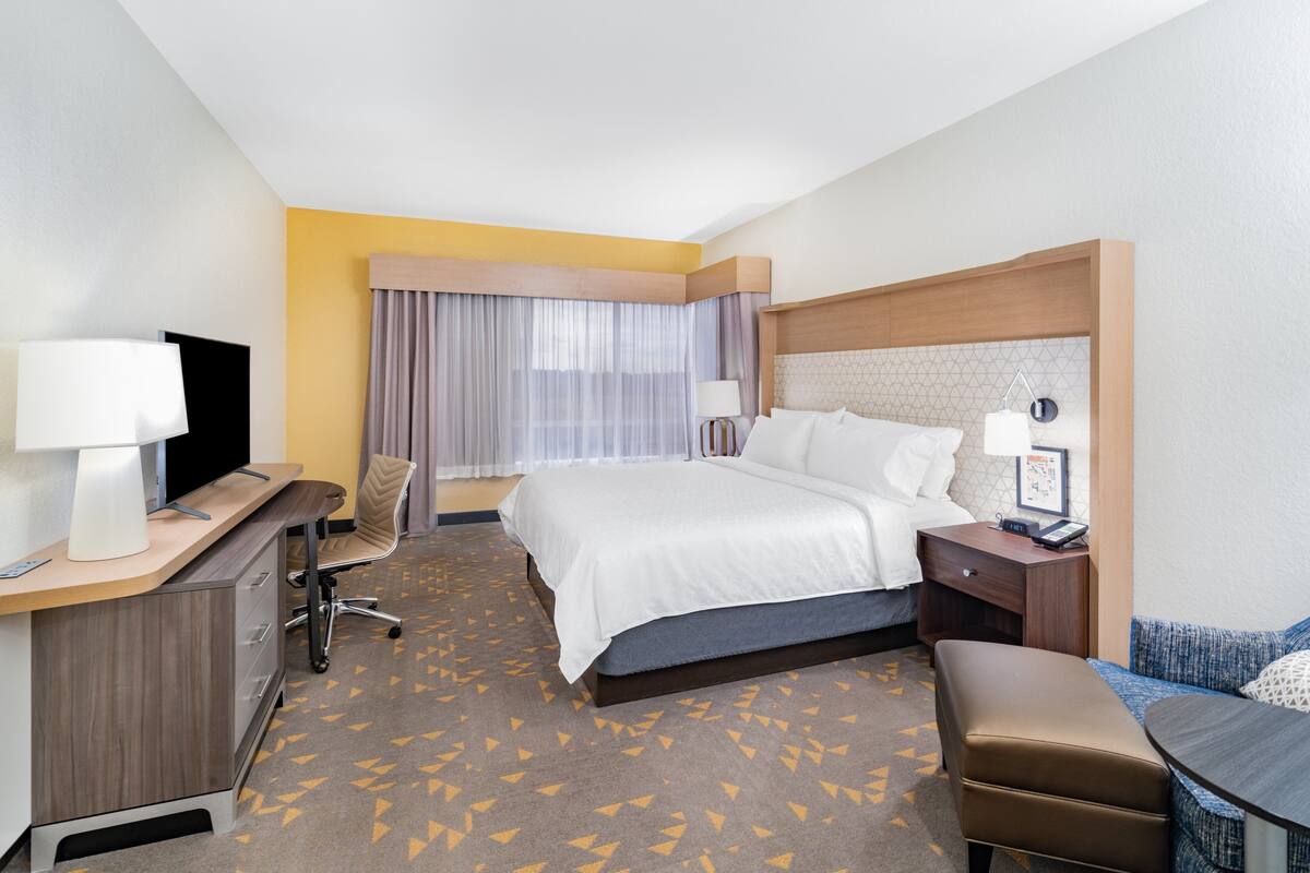 A hotel room with a bed, desk, chair, and a small table with a lamp.