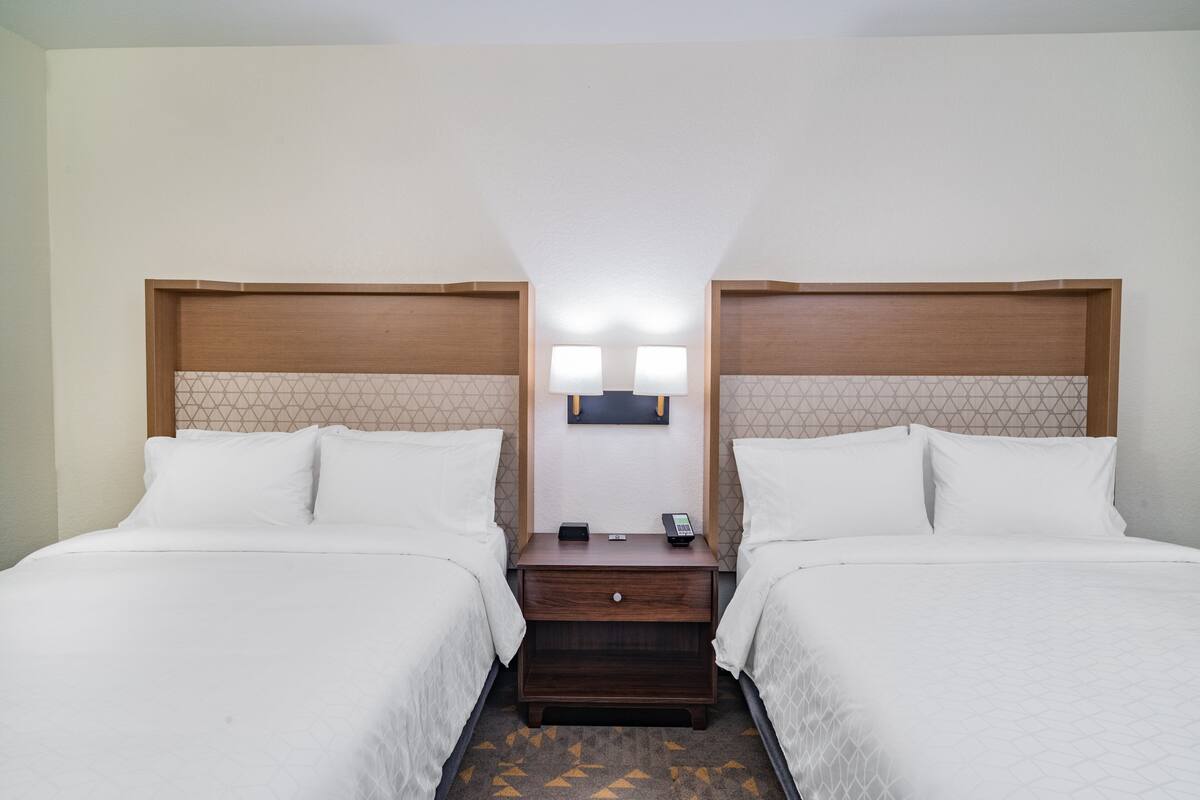 A hotel room with two beds, a nightstand with a lamp, and a small table.