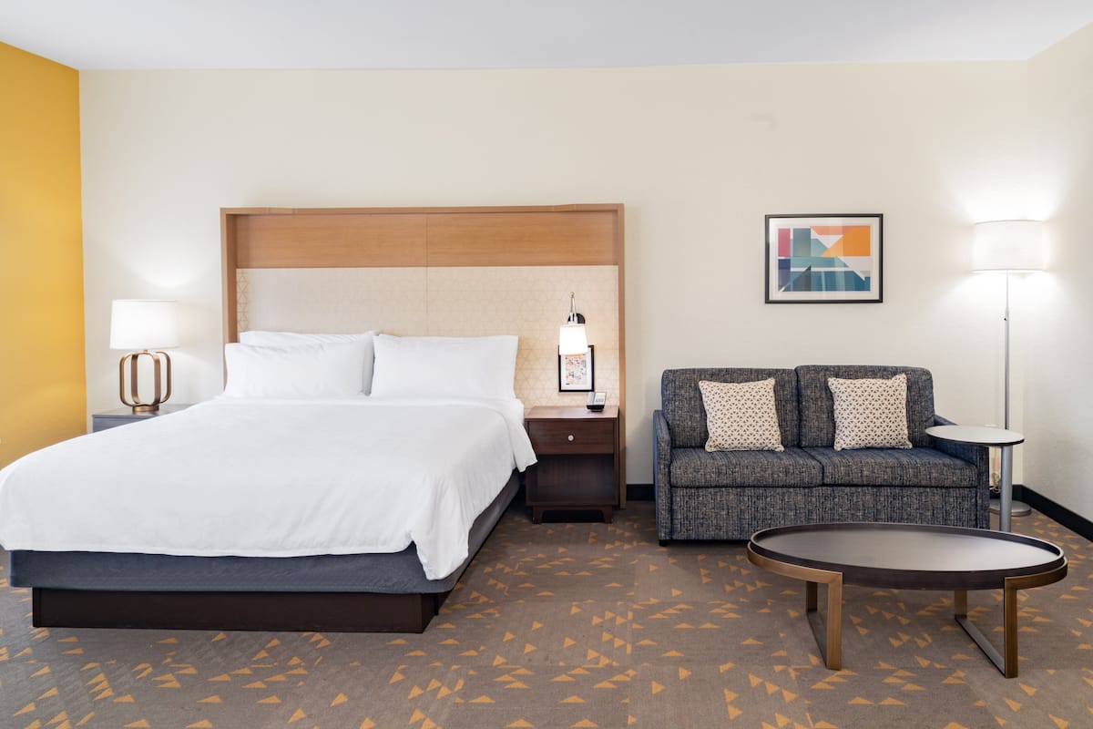 A hotel room with a bed, a sofa, a small table, a nightstand, and a lamp.