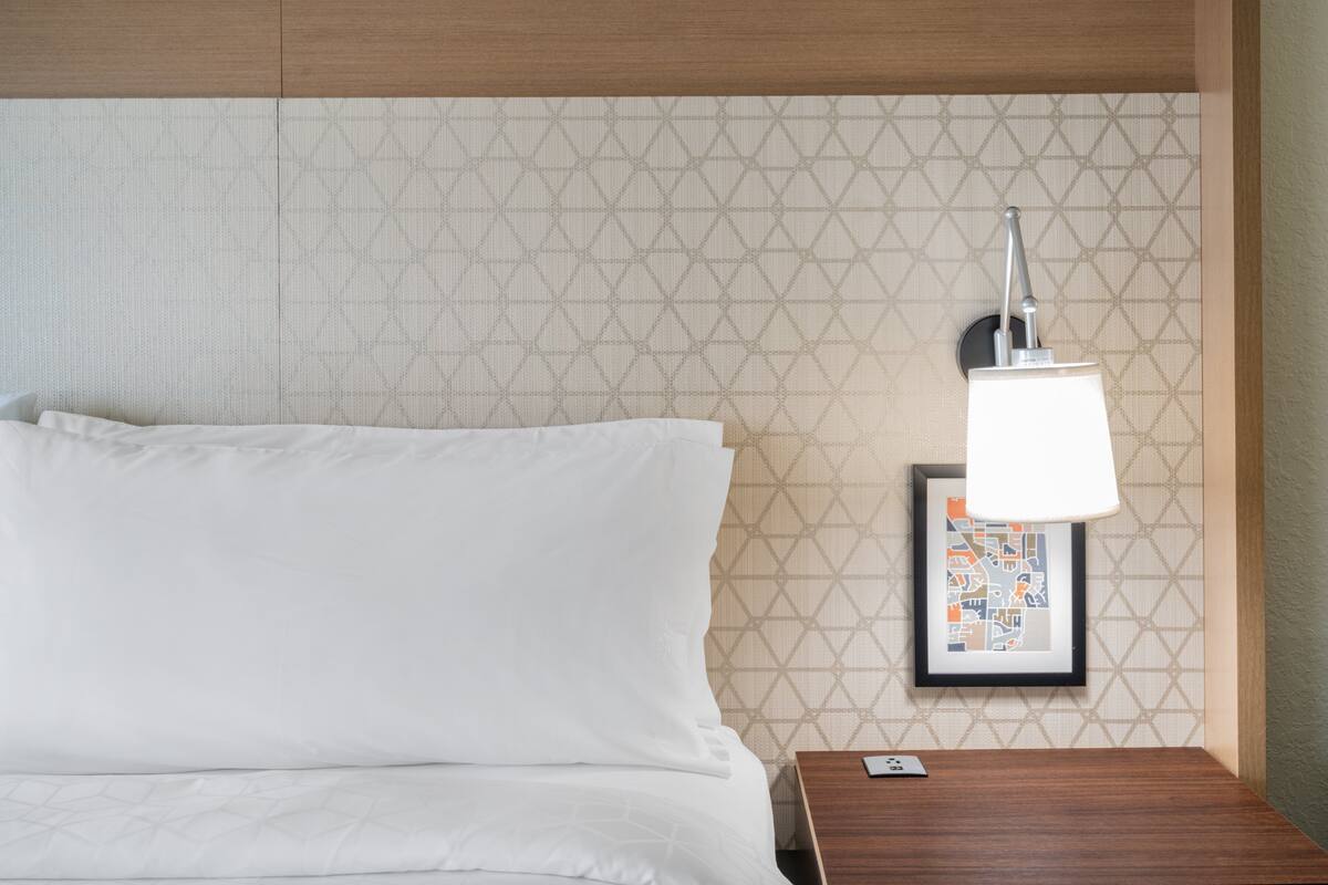 A neatly made bed with white linens, a wooden nightstand with a framed picture and a lamp, and a patterned wall.