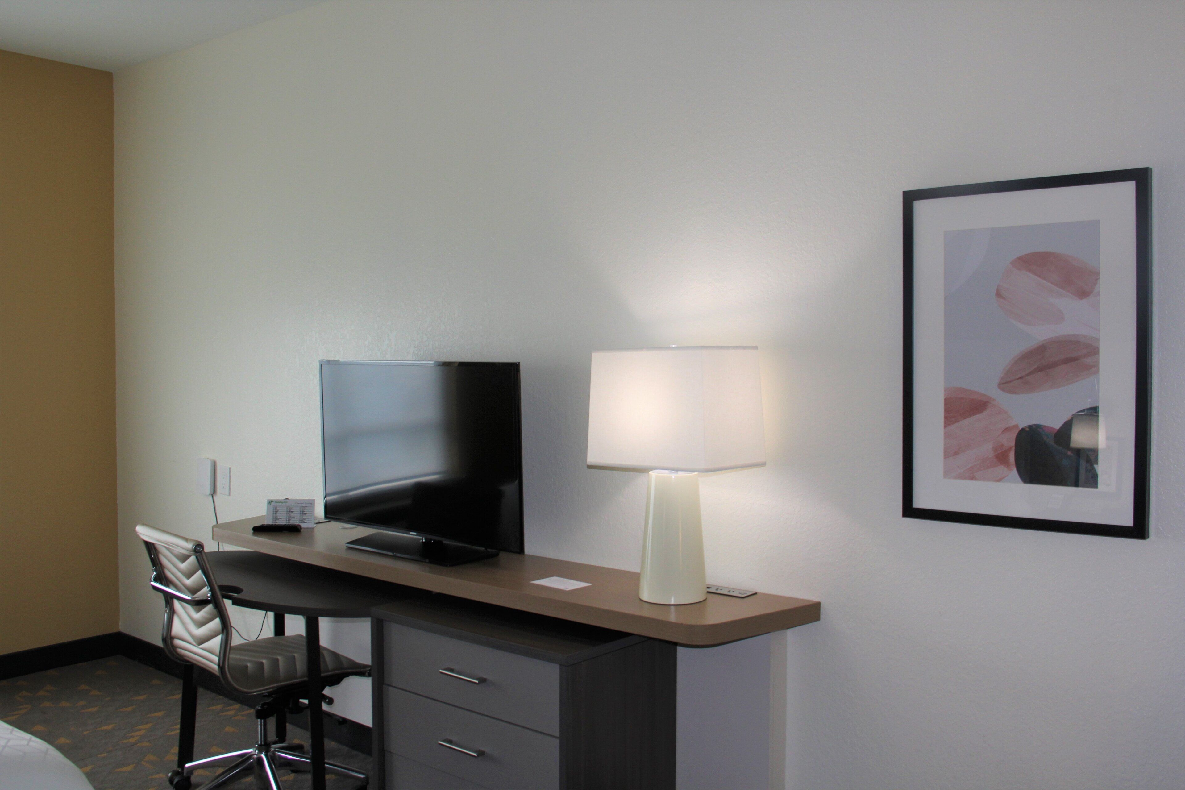 Ein Hotelzimmer mit Schreibtisch, Fernseher, Lampe, Stuhl und einem gerahmten Bild an der Wand.