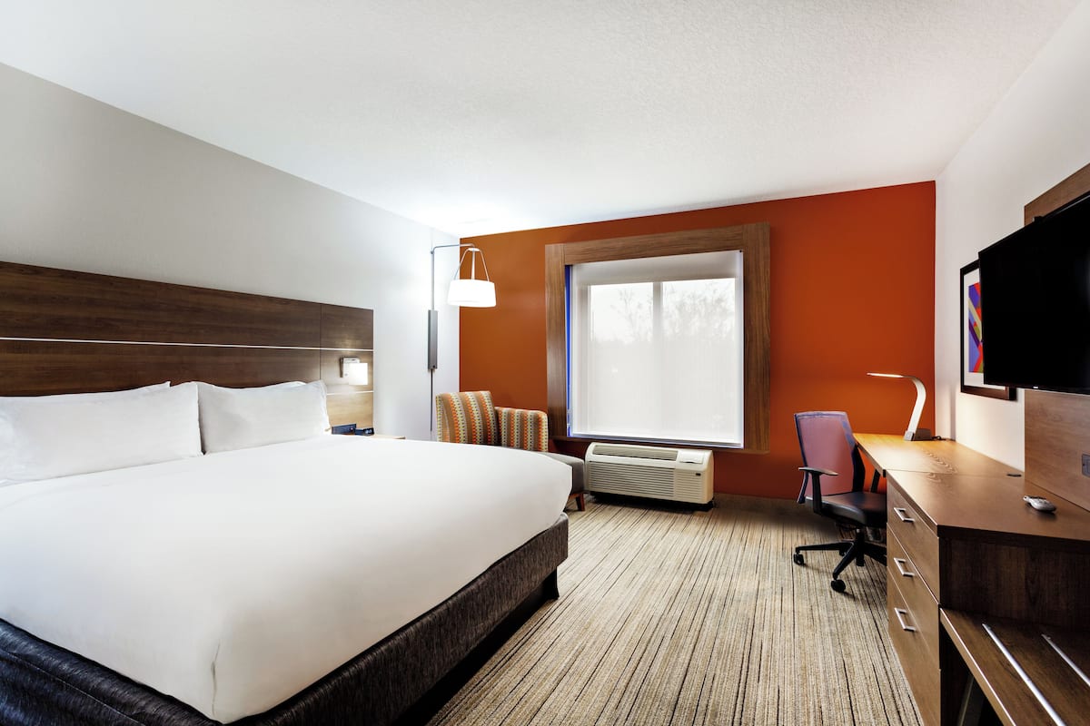 Une chambre d’hôtel avec un grand lit, un bureau, une chaise, une télévision et une fenêtre avec des stores.