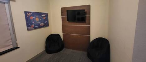 Quarto com parede de painel de madeira, televisão montada, dois puffs pretos e um pôster com tema espacial na parede.