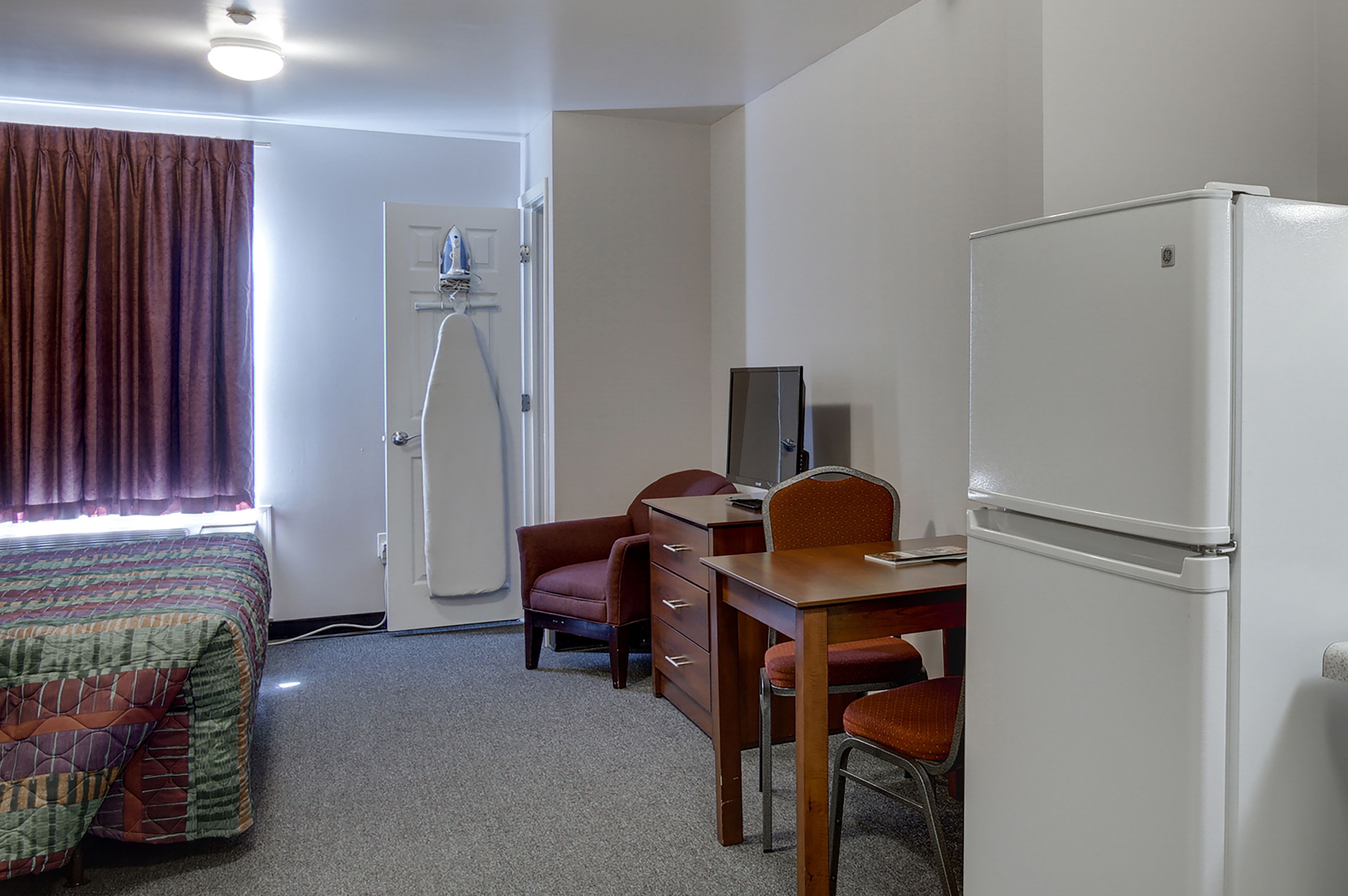 A hotel room with a bed, a desk with a chair, a refrigerator, and a shower.
