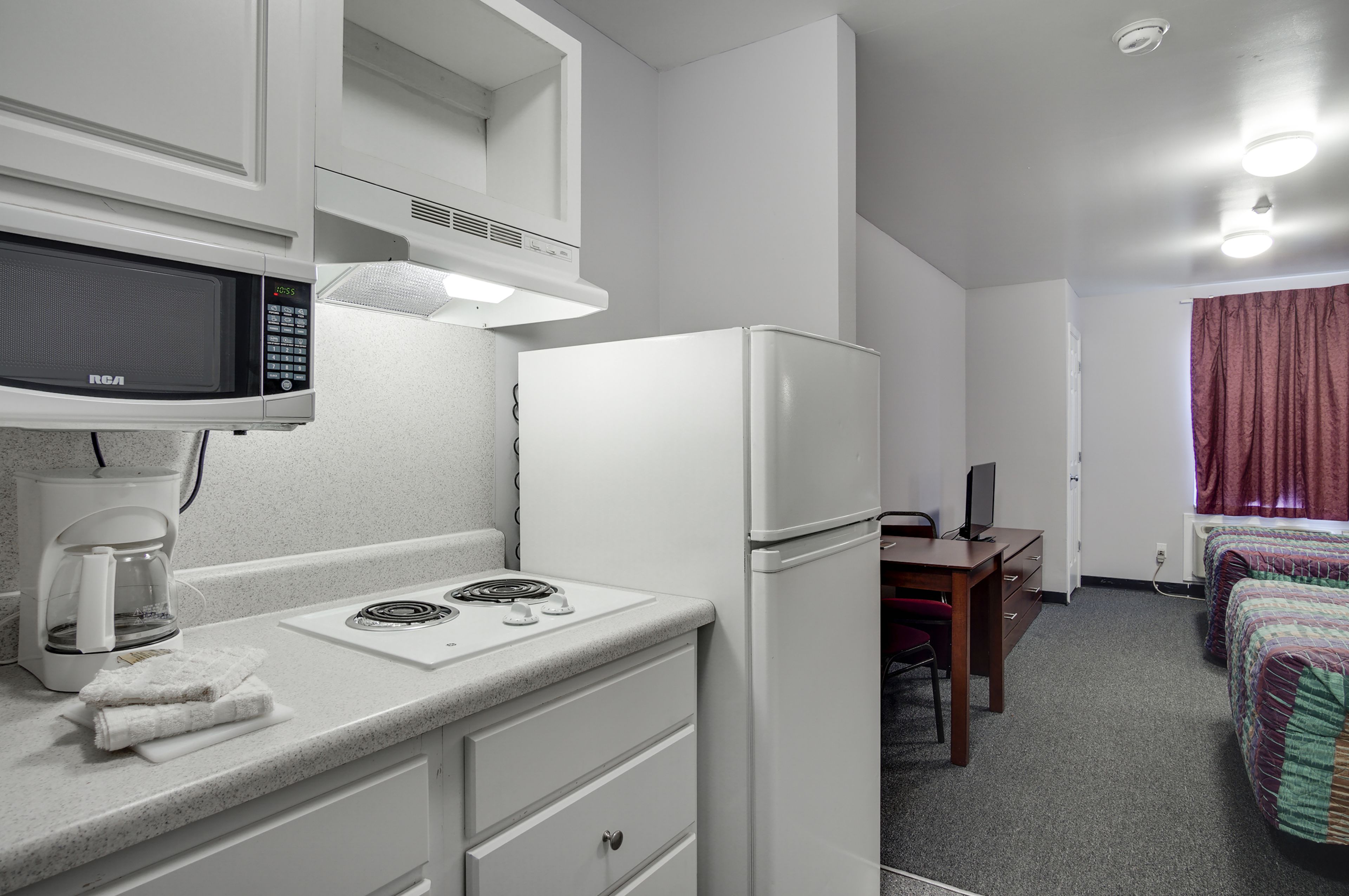 Una kitchenette compacta con microondas, cafetera y refrigerador. Hay un pequeño comedor con una mesa y una silla, y una cama con una manta colorida.