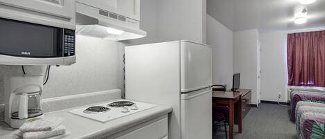 Una kitchenette compacta con microondas, cafetera y refrigerador. Hay un pequeño comedor con una mesa y una silla, y una cama con una manta colorida.