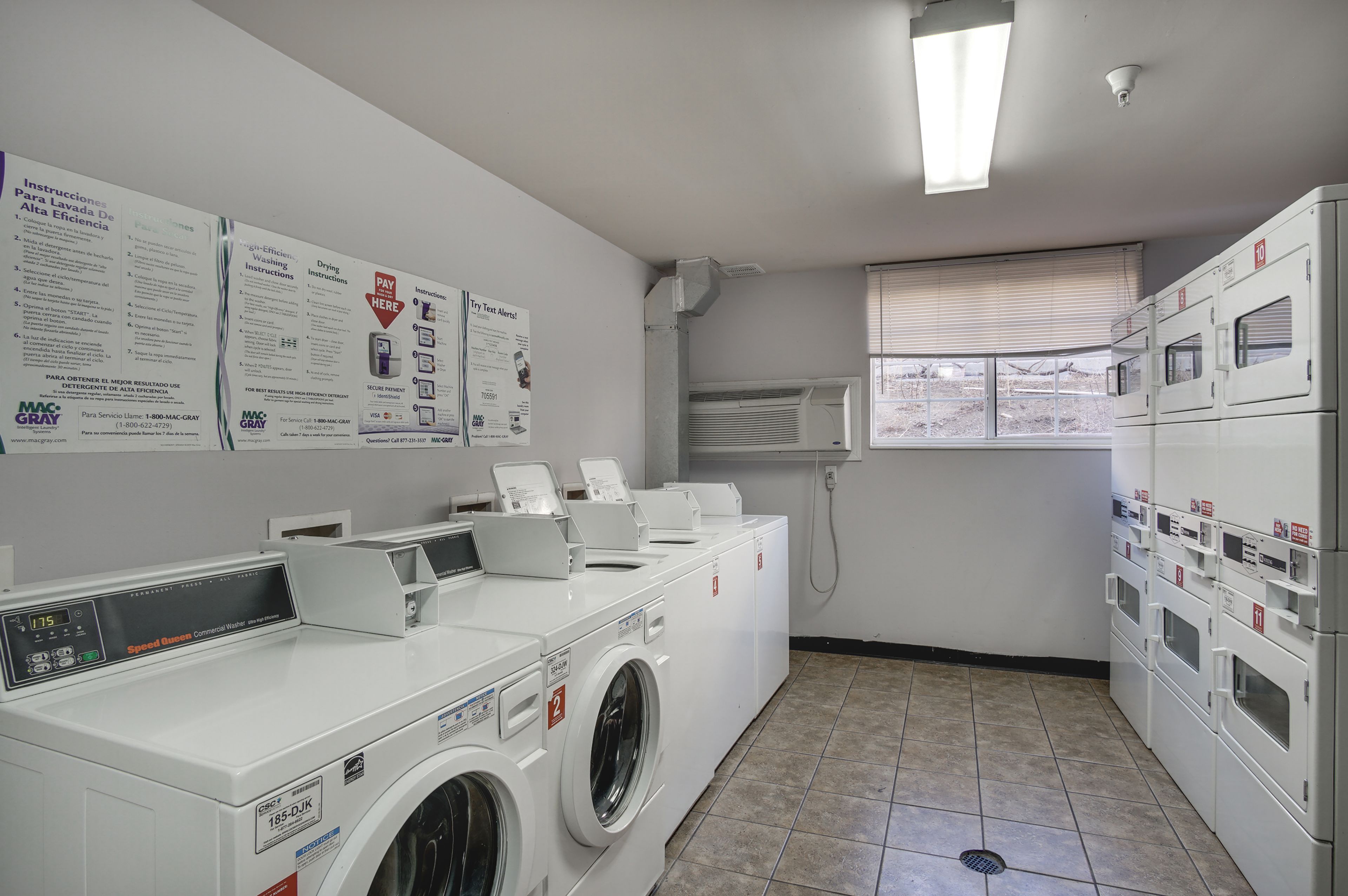 A laundry facility with multiple washing machines and dryers, instructional posters on the wall, and a window with blinds.