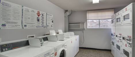 A laundry facility with multiple washing machines and dryers, instructional posters on the wall, and a window with blinds.