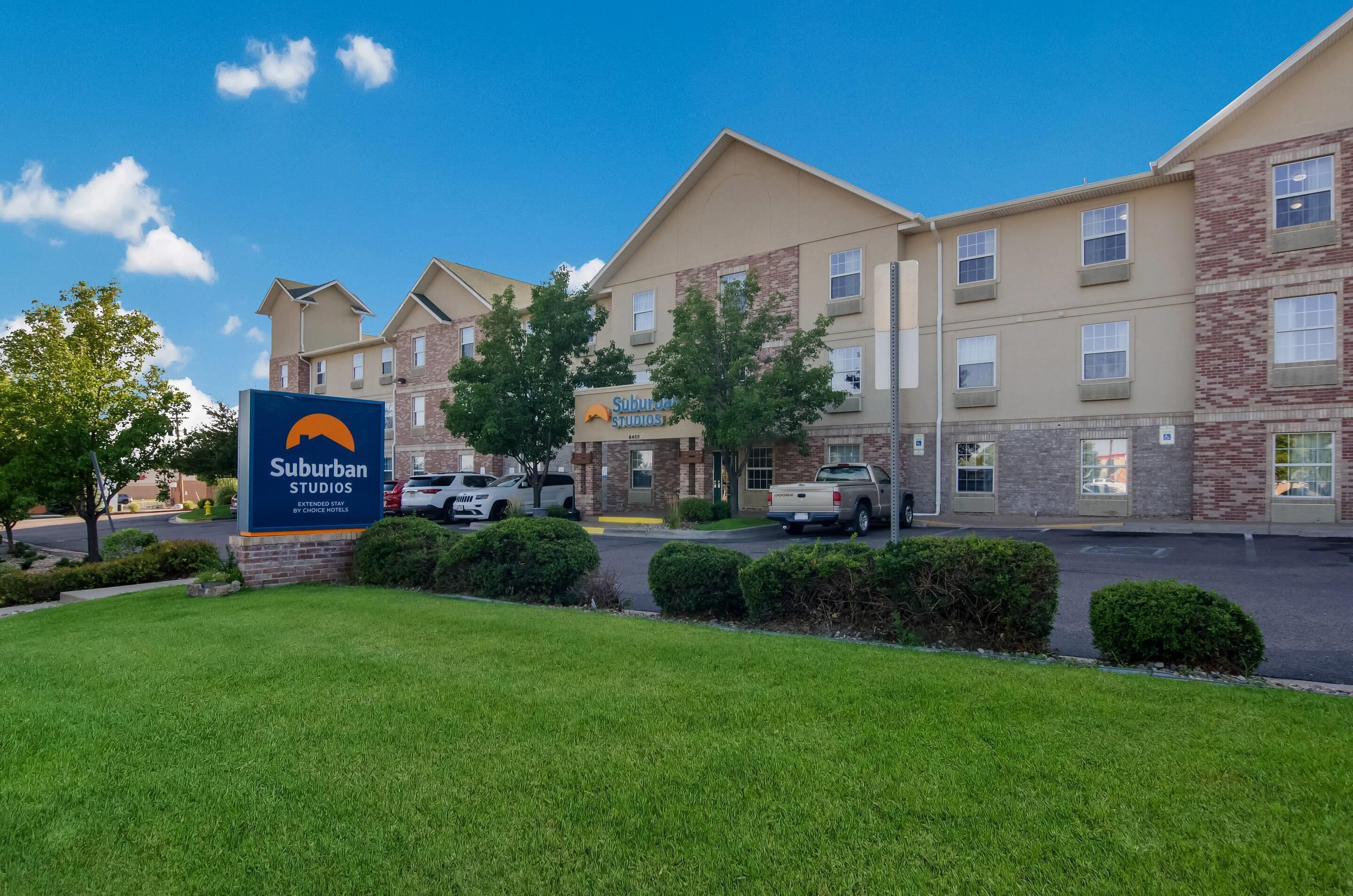 A Suburban Studios hotel with a sign, parking lot, and buildings with brick and stone facades.