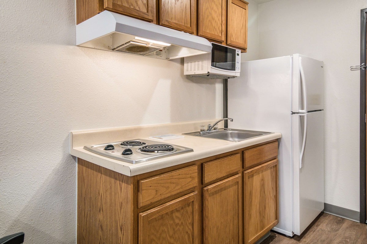 A compact kitchen with wooden cabinets, a built-in microwave, a stainless steel sink, and a white refrigerator.