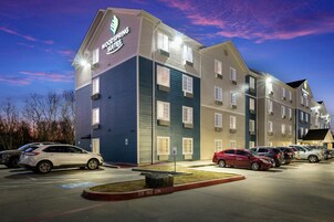 Un hotel de varios pisos con un cartel que dice 'Woodspring Suites'. Hay varios autos estacionados en la parte delantera, y se ve un letrero que indica 'Zona de remolque'.