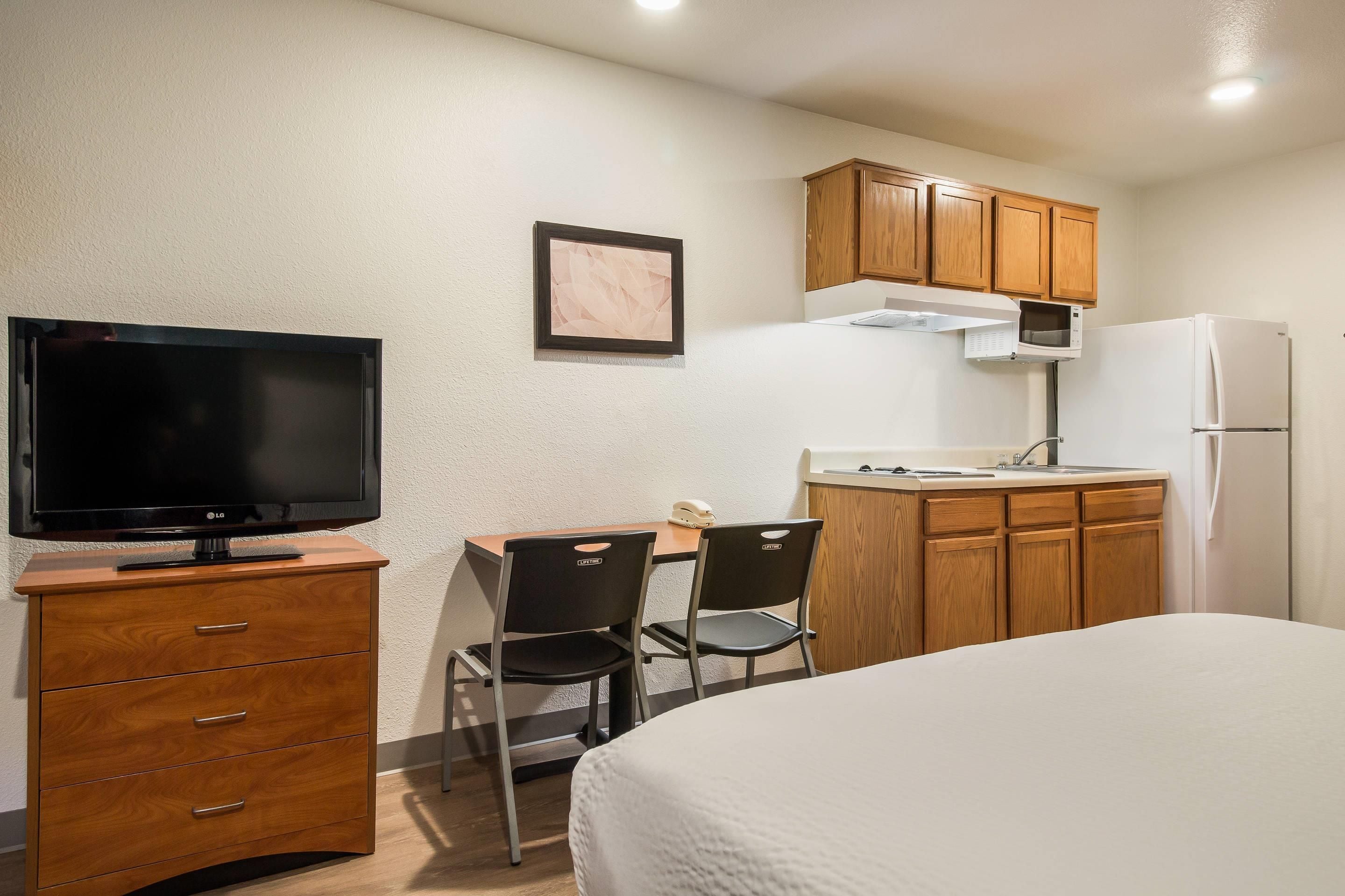 Quarto de hotel pequeno, com cama, televisão, cozinha compacta com micro-ondas e frigorífico, armário de madeira e escrivaninha com duas cadeiras.