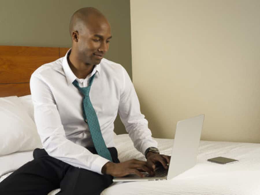 A man in a white shirt and teal tie using a laptop on a bed with white bedding and a wooden headboard.