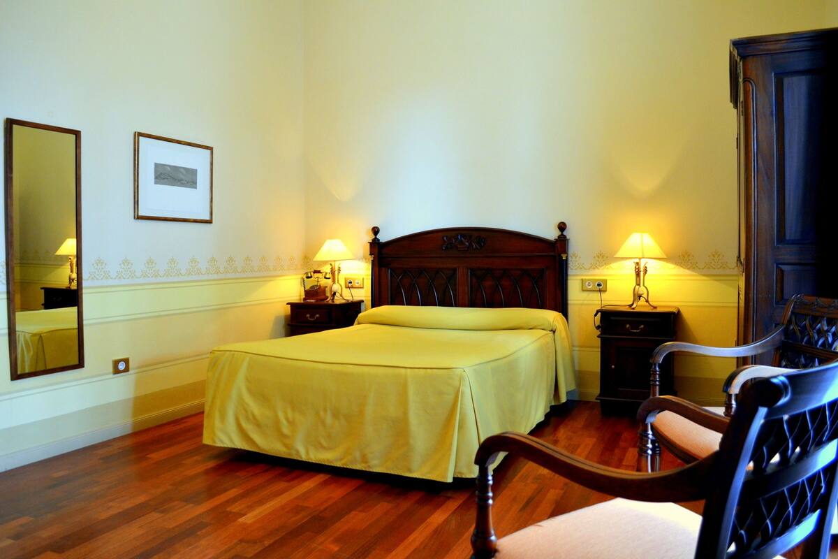 A bedroom with a wooden bed, two bedside tables with lamps, a chair, a mirror, and a framed picture on the wall.