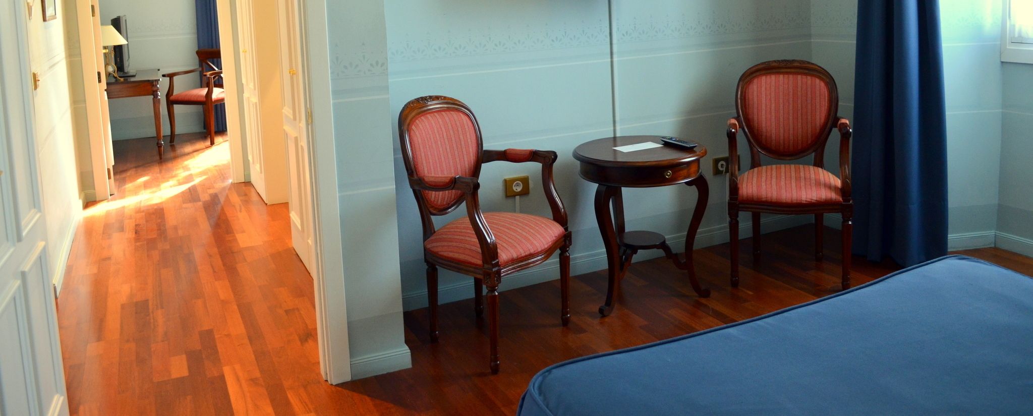 A hotel room with a wooden floor, two upholstered chairs, a small round table, and a bed with a blue cover.