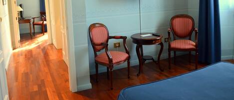 A hotel room with a wooden floor, two upholstered chairs, a small round table, and a bed with a blue cover.